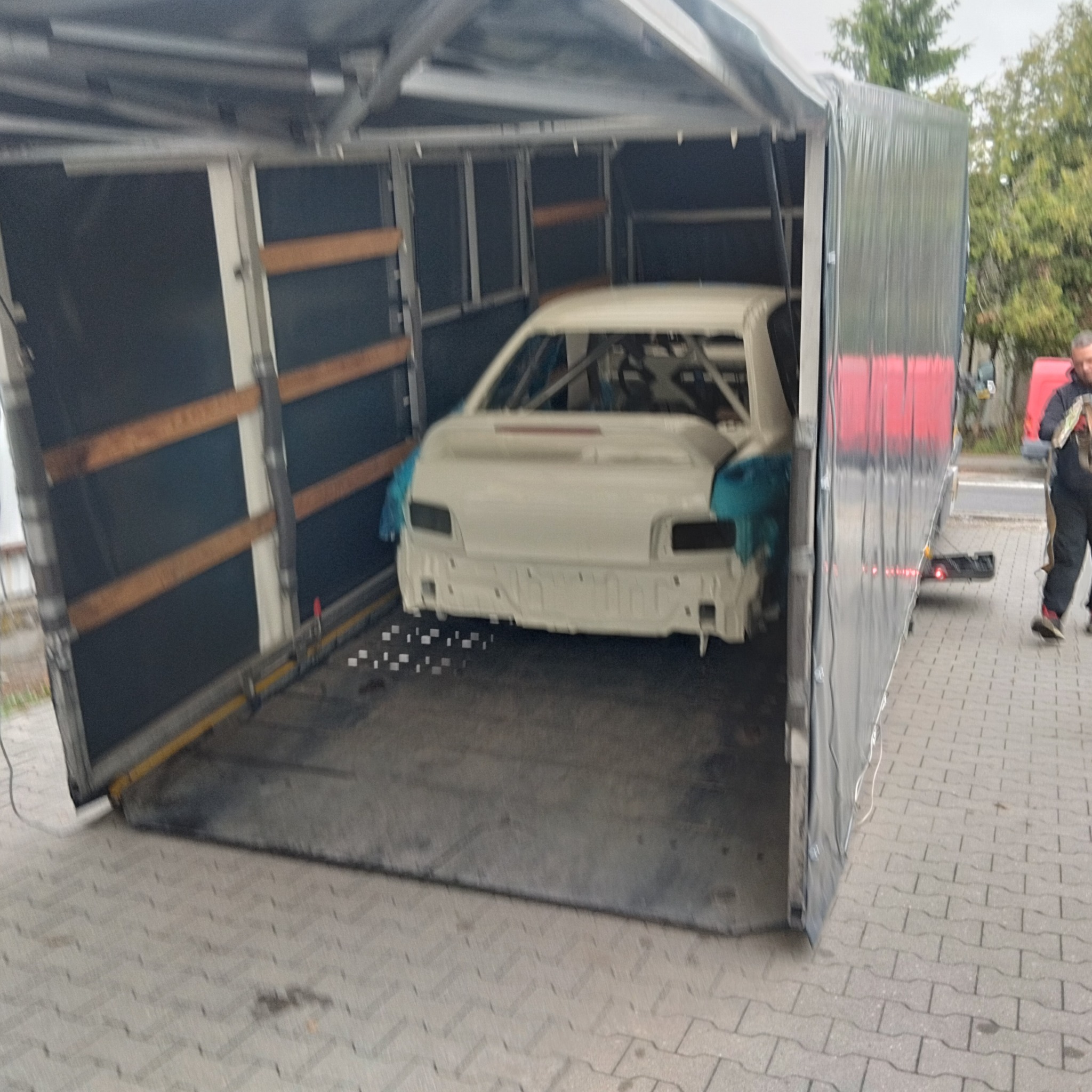 Transport samochodu wyścigowego w specjalistycznej zabudowie lawety. Biały pojazd z klatką bezpieczeństwa. Widoczna osoba przy lawetcie.