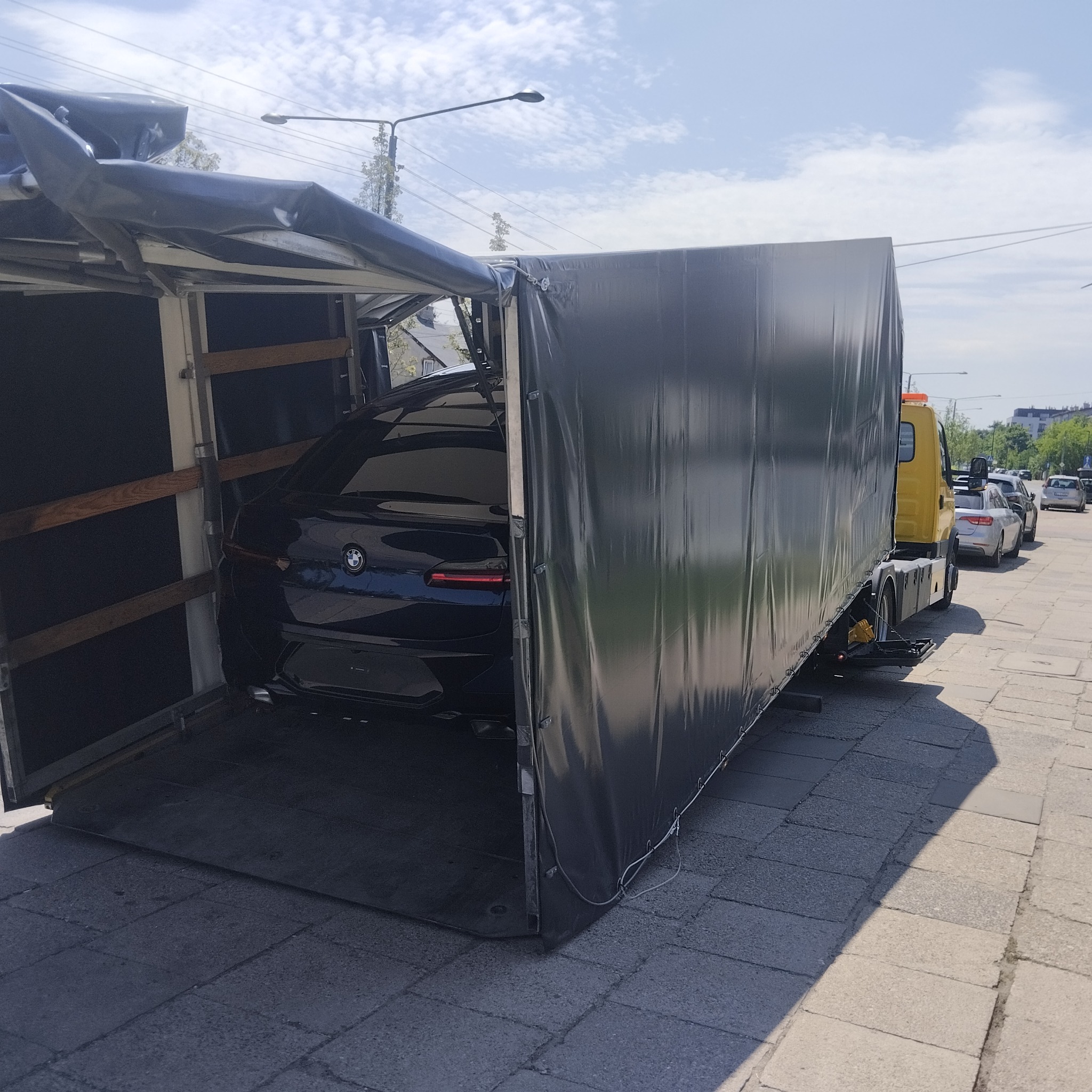 Czarny samochód BMW na platformie załadunkowej lawety z czarną plandeką, widok z tyłu pojazdu i boku lawety na tle miejskiej ulicy.