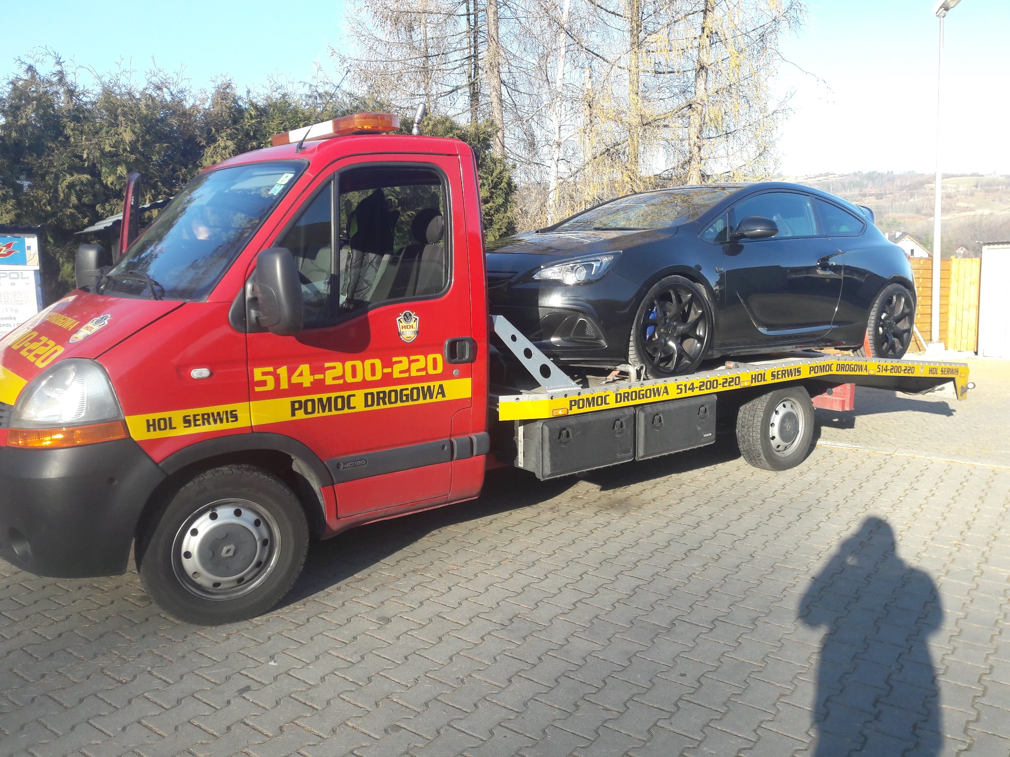 Czerwona autolaweta z czarnym Oplem Astrą GTC na platformie, zaparkowana w Pruszkowie na brukowanym placu w słoneczny dzień. Na pojeździe widoczne numery telefonu i napis 'Pomoc Drogowa'.