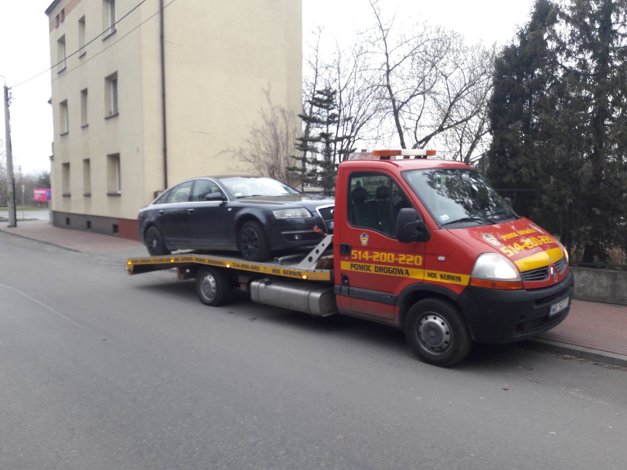 Ciemny samochód osobowy marki Audi transportowany na czerwonej ciężarówce pomocy drogowej z numerem telefonu i napisem 'Pomoc Drogowa' na tle budynku i drzew.