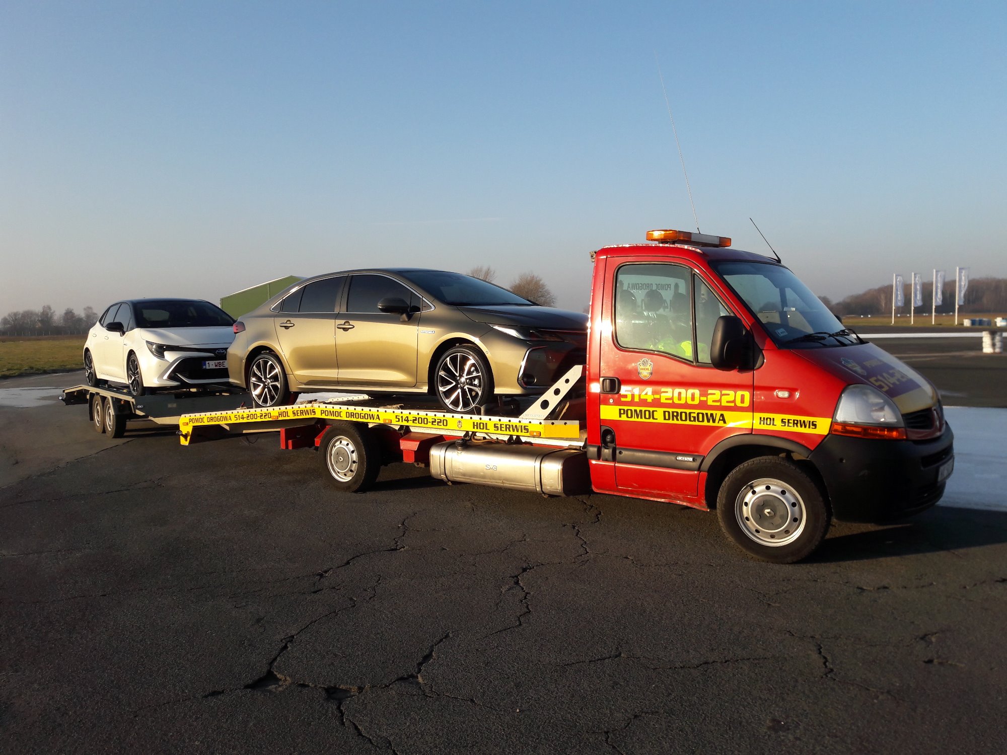 Czerwony pojazd pomocy drogowej z dwoma samochodami osobowymi na platformie, widok z boku, na tle lotniska w słoneczny dzień.