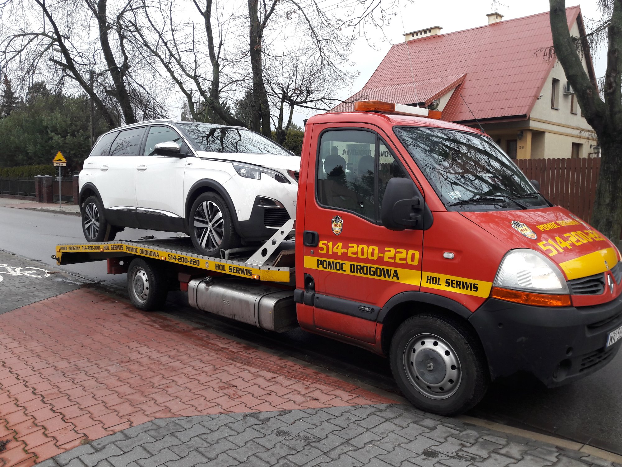 Czerwona ciężarówka pomocy drogowej z białym SUV-em na platformie, zaparkowana na chodniku z czerwonej kostki, w tle dom z czerwonym dachem.