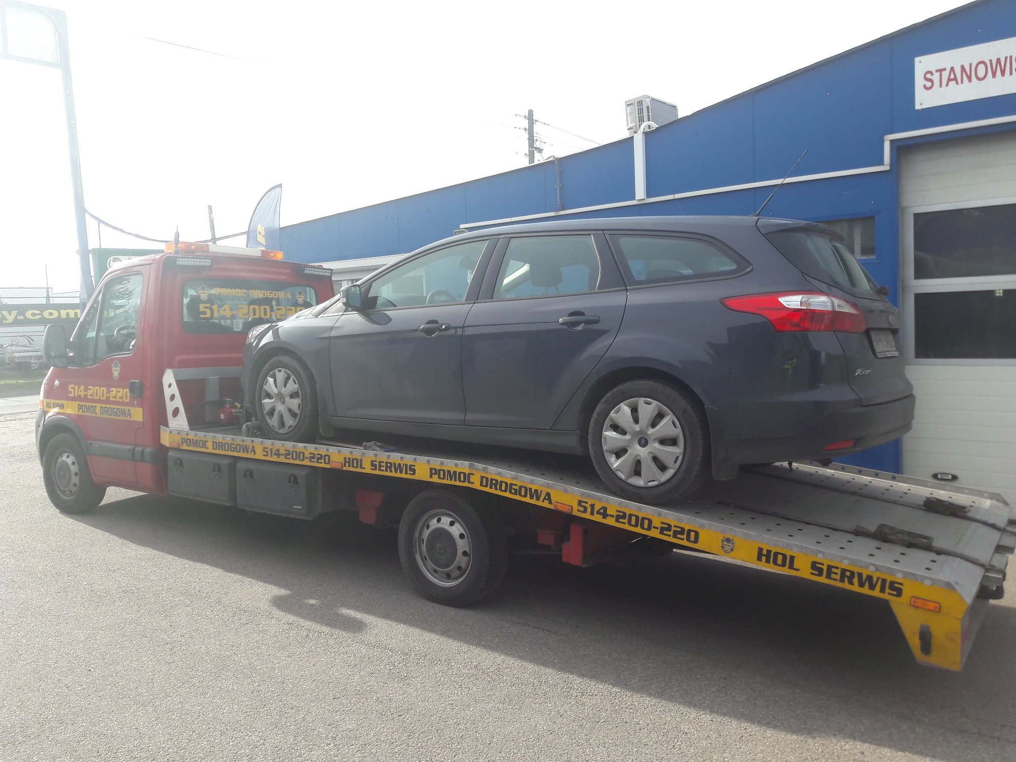 Ciemny samochód kombi marki Ford Focus transportowany na czerwono-żółtej platformie pojazdu pomocy drogowej z widocznym numerem telefonu. W tle błękitny budynek warsztatu.