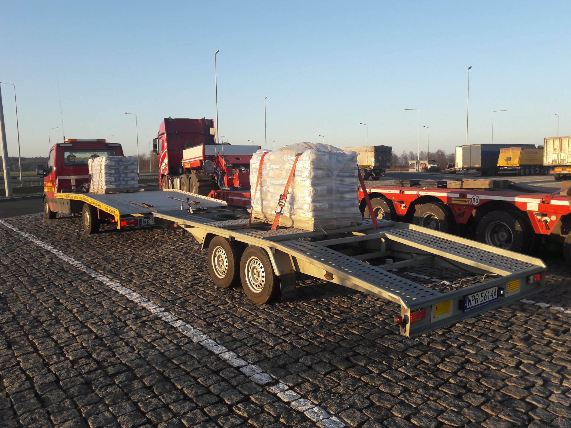 Czerwona ciężarówka z platformą transportową przewożąca paletę owiniętą folią, zaparkowana na brukowanym parkingu w słoneczny dzień.