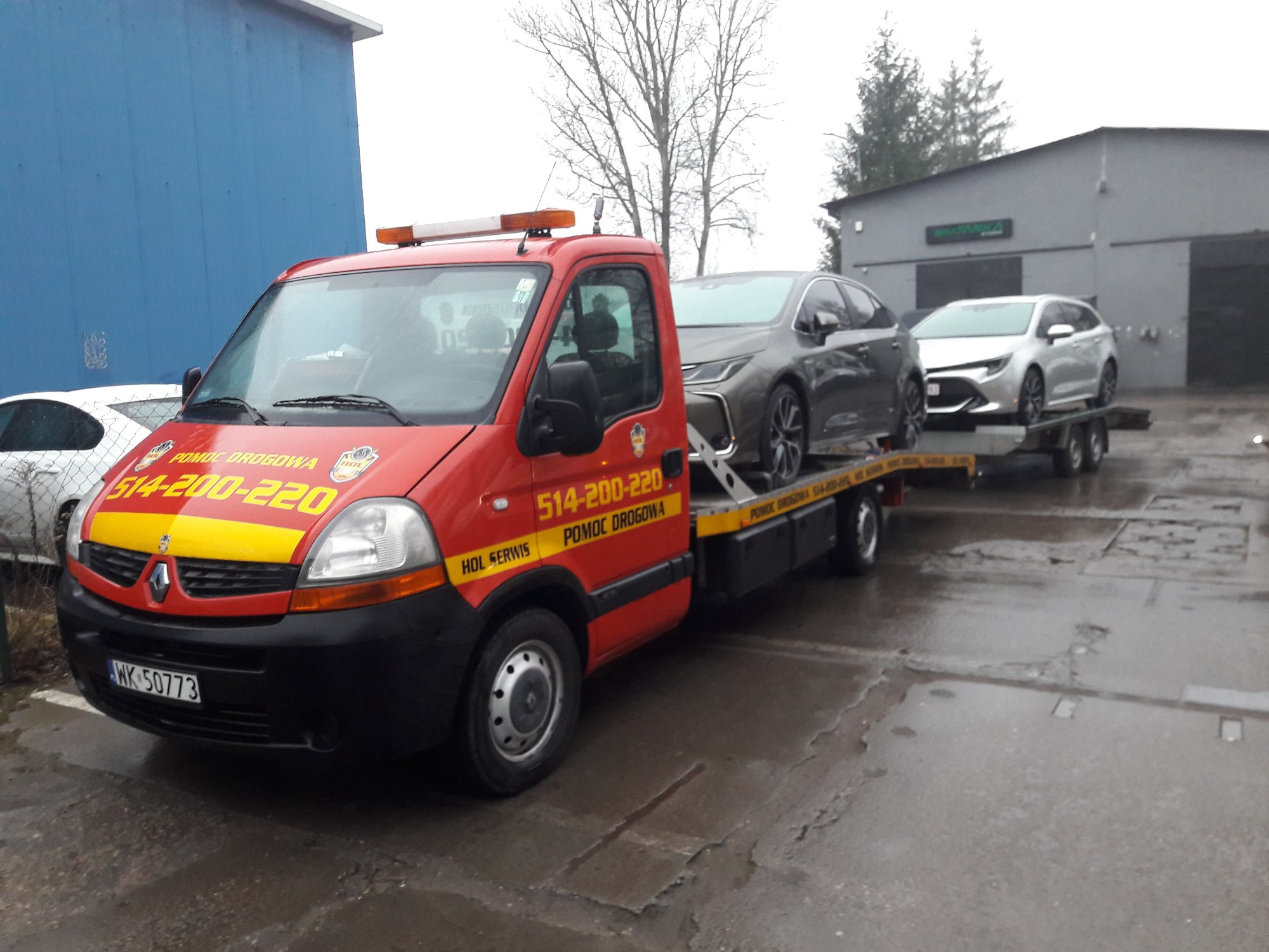 Czerwona ciężarówka pomocy drogowej z dwoma samochodami osobowymi na platformie, zaparkowana na mokrym placu przed budynkiem z niebieską ścianą.