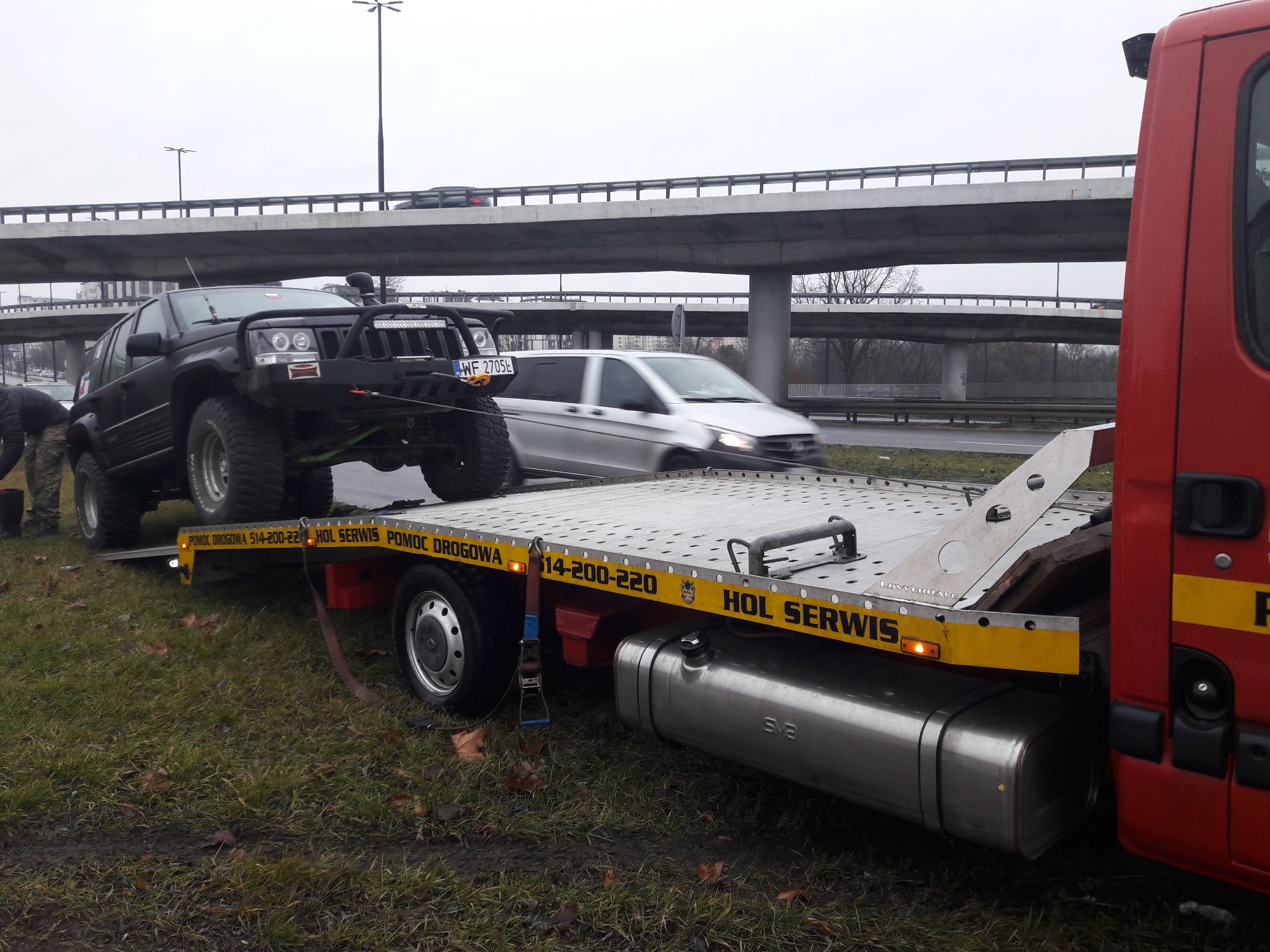 Czarny terenowy Jeep z dodatkowym oświetleniem LED na platformie pomocy drogowej z widocznym numerem rejestracyjnym WF 2705L, na tle wiaduktu i przemieszczających się pojazdów.