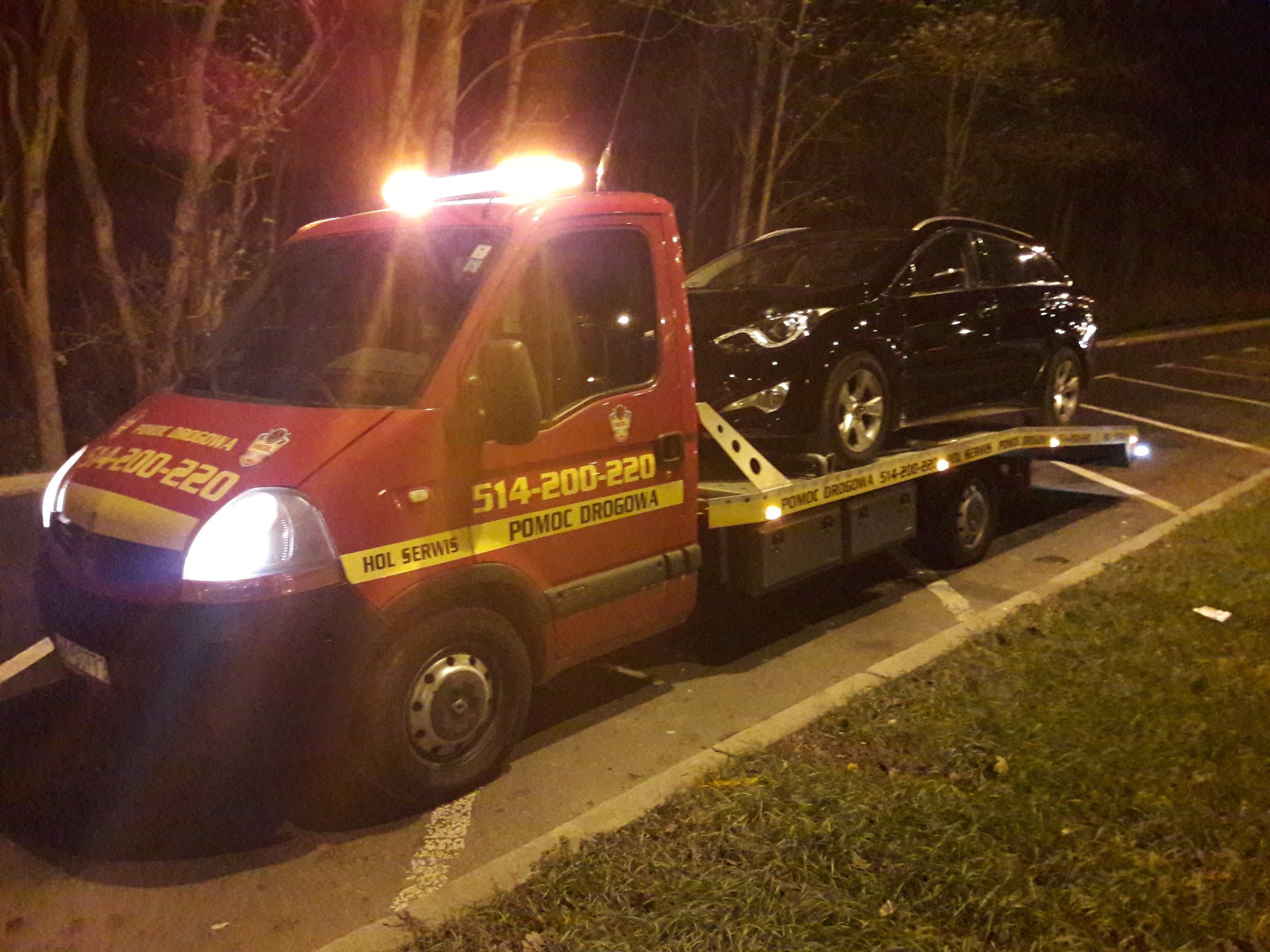 Czerwony pojazd pomocy drogowej z włączonymi światłami ostrzegawczymi przewozi czarny samochód osobowy na platformie, widoczne numery kontaktowe i logo firmy na boku pojazdu, nocne ujęcie