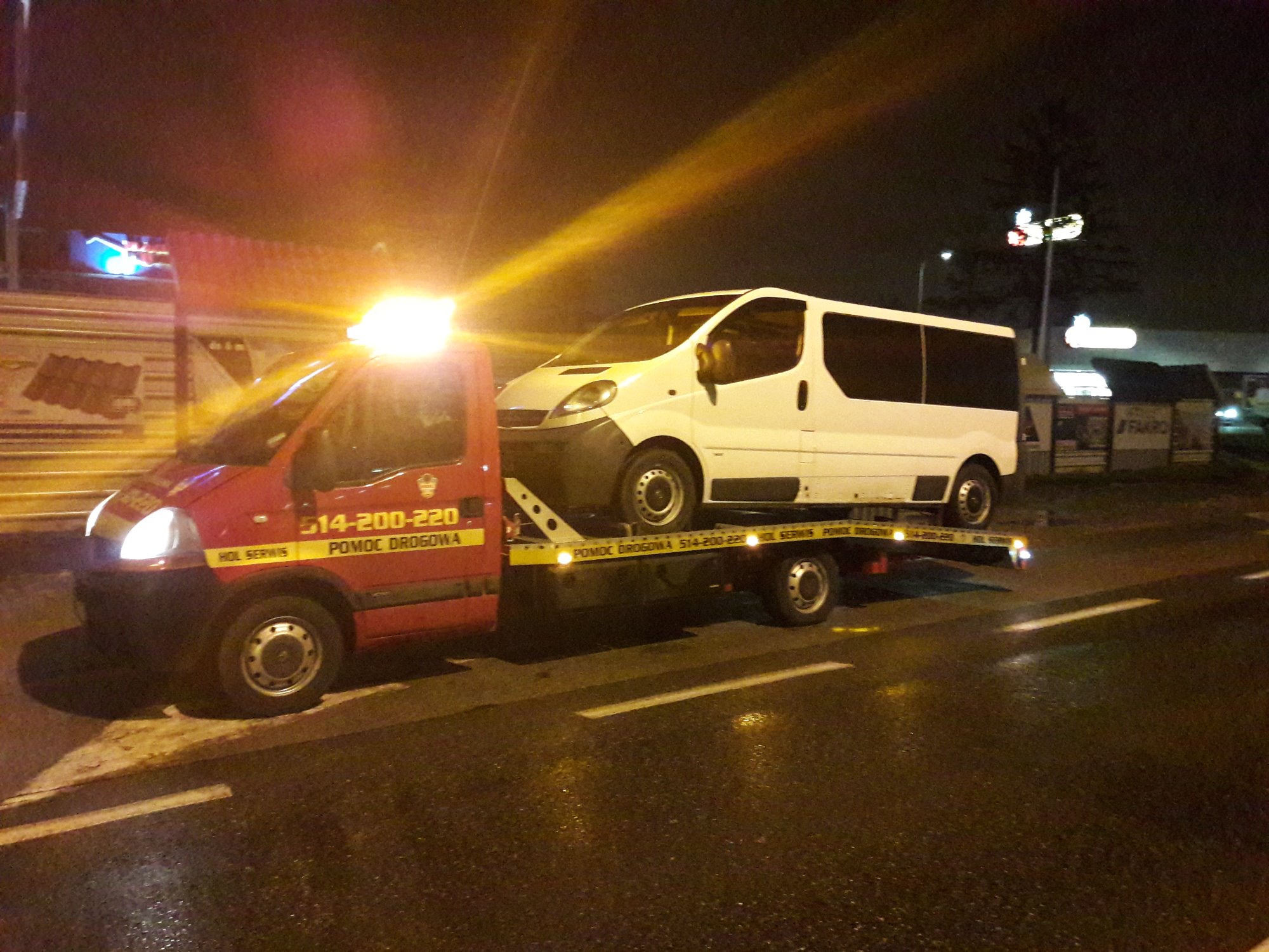 Czerwona ciężarówka pomocy drogowej z włączonymi światłami ostrzegawczymi transportuje biały samochód dostawczy na platformie w nocy. Na karoserii ciężarówki widoczny numer telefonu.
