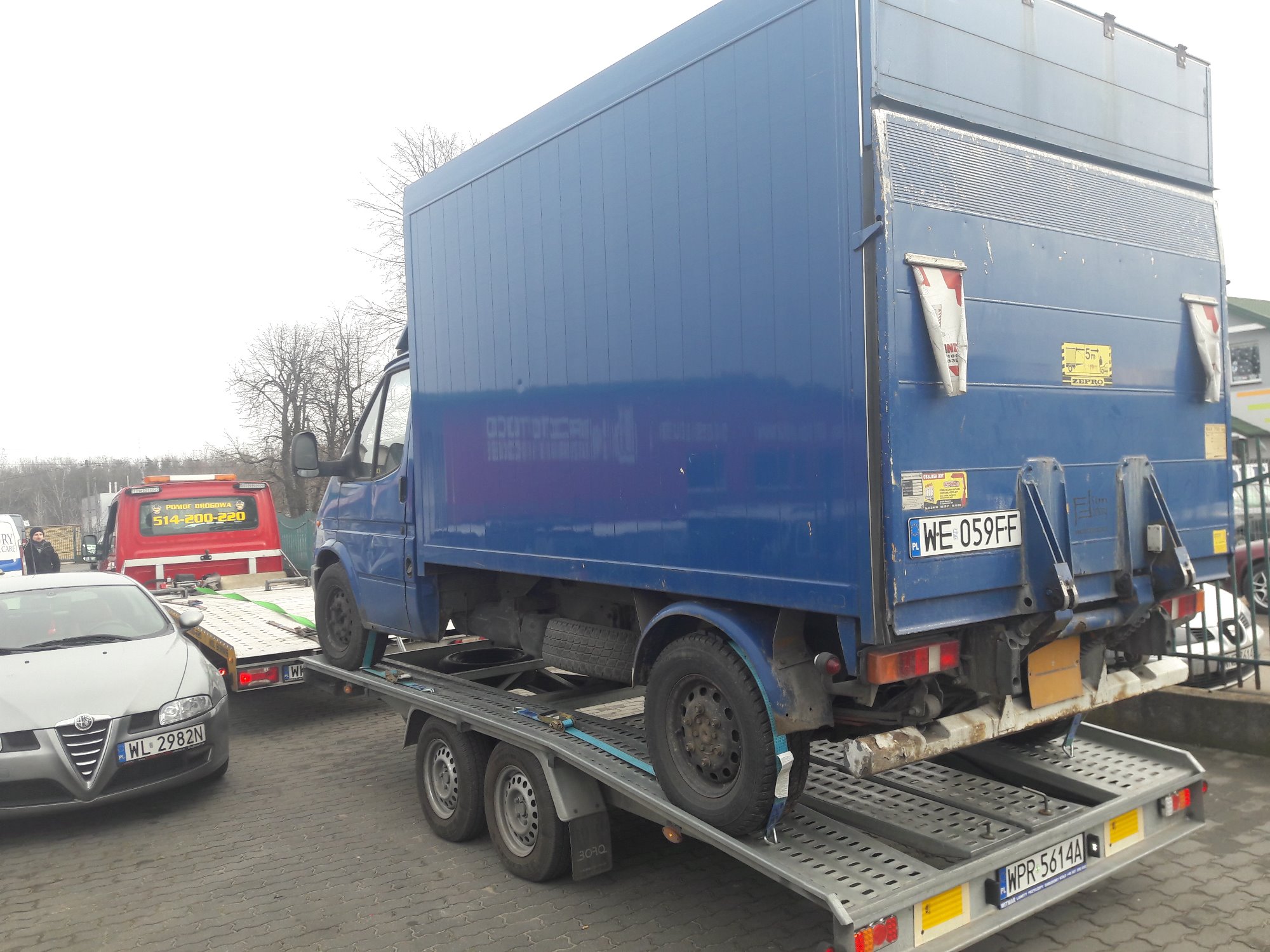 Laweta transportuje niebieski samochód dostawczy z zabudową kontenerową, zabezpieczony niebieskimi pasami transportowymi. W tle widoczny czerwony samochód pomocy drogowej i srebrny samochód osobowy...