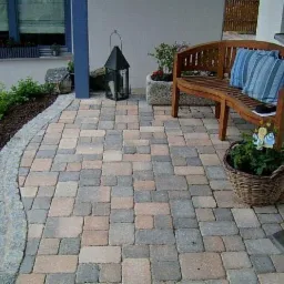 Ułożona kostka brukowa w odcieniach szarości i brązu, z zaokrąglonym brzegiem wykończonym kostką granitową, uzupełniona zielenią, ławką z poduszkami i dekoracyjną latarnią.