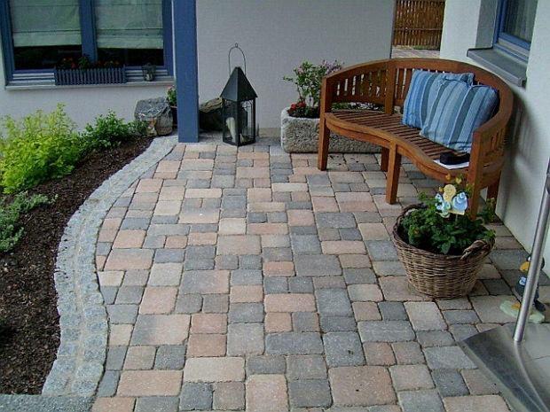 Ułożona kostka brukowa w odcieniach szarości i brązu, z zaokrąglonym brzegiem wykończonym kostką granitową, uzupełniona zielenią, ławką z poduszkami i dekoracyjną latarnią.