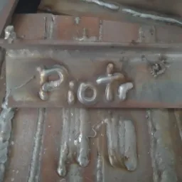 Spawy na metalu układające się w imię 'Piotr', z widocznymi śladami obróbki cieplnej i nierównościami spoin. Surowy, industrialny charakter.