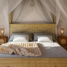 Jasne drewniane łóżko z baldachimem i grubym, dzierganym pledem. Lampki nocne z ryflowanymi kloszami. Ujęcie centralne, spokojna kolorystyka.