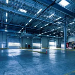 Wnętrze hali magazynowej z oświetleniem przemysłowym, widoczne bramy rolowane i ciężarówka z ładunkiem samochodów.