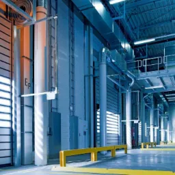 Wnętrze hali magazynowej z instalacją elektryczną: widoczne rury, kable, oświetlenie sufitowe i brama segmentowa z oznaczeniem 'E.p', w tle zaparkowana ciężarówka.