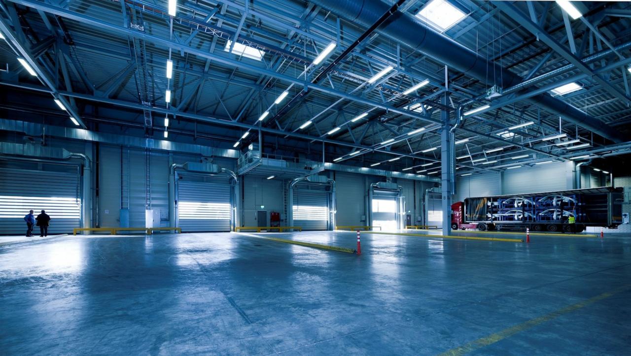 Wnętrze hali magazynowej z oświetleniem przemysłowym, widoczne bramy rolowane i ciężarówka z ładunkiem samochodów.