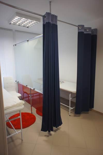 Gabinet zabiegowy z zasłonami oddzielającymi stanowiska, widoczny stolik zabiegowy i krzesła, oświetlenie sufitowe.