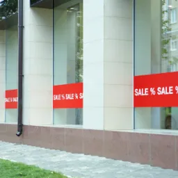 Elewacja budynku z oknami, na których umieszczono czerwone banery z napisem 'SALE %' w białej czcionce, widoczne odbicie otoczenia w szybach.