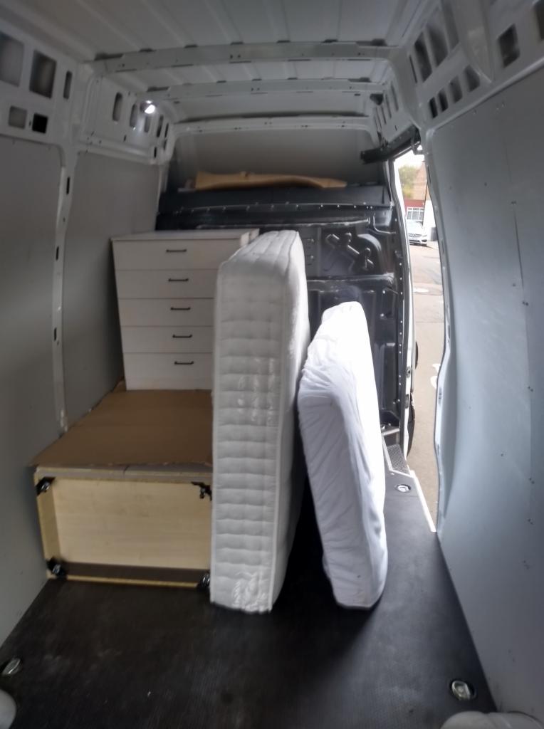 Wnętrze furgonetki załadowane komodą z szufladami, kartonowym pudłem i dwoma materacami owiniętymi folią, przygotowanymi do transportu.