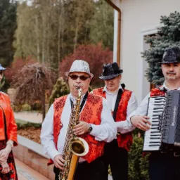 Czteroosobowy zespół muzyczny w czerwono-czarnych kamizelkach i kapeluszach grający na saksofonie, akordeonie i tamburynie na tle zieleni.