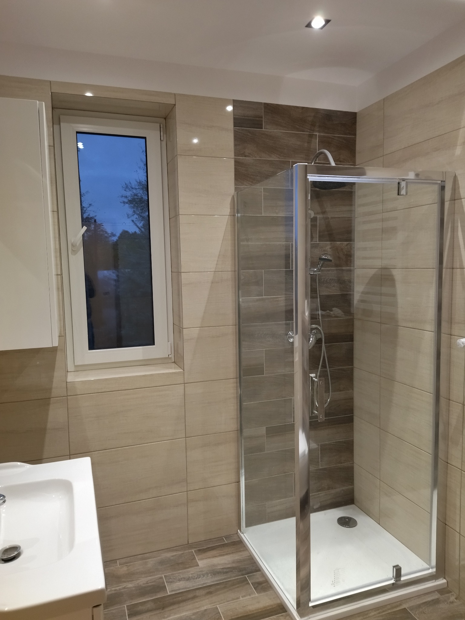 Nowoczesna łazienka z kabiną prysznicową typu walk-in, wyłożona beżowymi i brązowymi płytkami imitującymi drewno, z widoczną umywalką i oknem z widokiem na zewnątrz.
