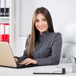 Uśmiechnięta kobieta w szarej koszuli siedzi przy biurku z laptopem. W tle białe biuro z segregatorami i roletami.