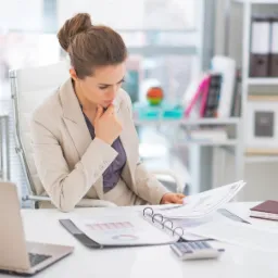 Kobieta w biurze analizuje dokumenty z wykresami, siedząc przy biurku z laptopem i kalkulatorem. W tle regał z segregatorami, jasne wnętrze.