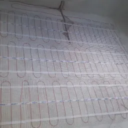 Rozłożona mata grzewcza DEVI z czerwonymi kablami na przygotowanym podłożu w trakcie instalacji ogrzewania podłogowego w pomieszczeniu.