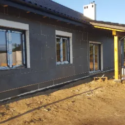 Dom w trakcie ocieplania elewacji, widoczne szare płyty styropianowe, okna z folią ochronną oraz drewniana konstrukcja zadaszenia tarasu.