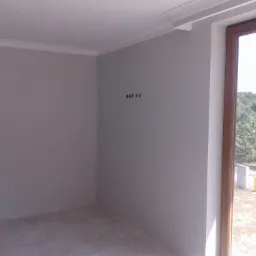 Pomalowane na szaro wnętrze pokoju z widocznymi puszkami elektrycznymi na ścianie i oknem z drewnianą ramą, widok na zewnątrz na drzewa i elementy konstrukcyjne.