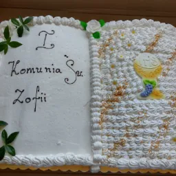 Prostokątny tort komunijny w kształcie otwartej księgi, z napisem 'I Komunia Św. Zofii' po lewej stronie i dekoracją kielicha z hostią oraz winogronami po prawej, całość zdobiona bitą śmietaną...