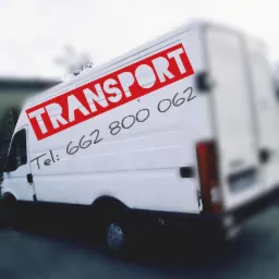 Biały bus z czerwonym napisem 'TRANSPORT' i numerem telefonu, widoczny z boku, lekko rozmazane tło.