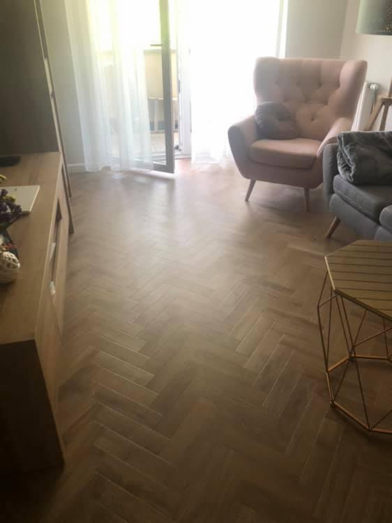 Parkiet ułożony w jodełkę francuską, ciepłe odcienie brązu, widoczne meble: różowy fotel typu uszak, szara sofa i stolik kawowy o geometrycznej konstrukcji, jasne wnętrze z oknem balkonowym.