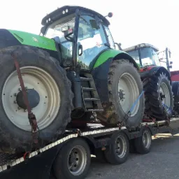 Dwa ciągniki rolnicze, jeden zielony, drugi czerwony, zabezpieczone pasami transportowymi na niskopodwoziowej naczepie ciężarówki, widok z boku na koła i kabiny.