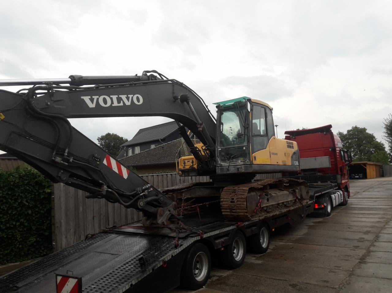 Żółto-czarna koparka Volvo na niskopodwoziowej naczepie ciężarówki, przygotowana do transportu, z widocznym domem w tle.