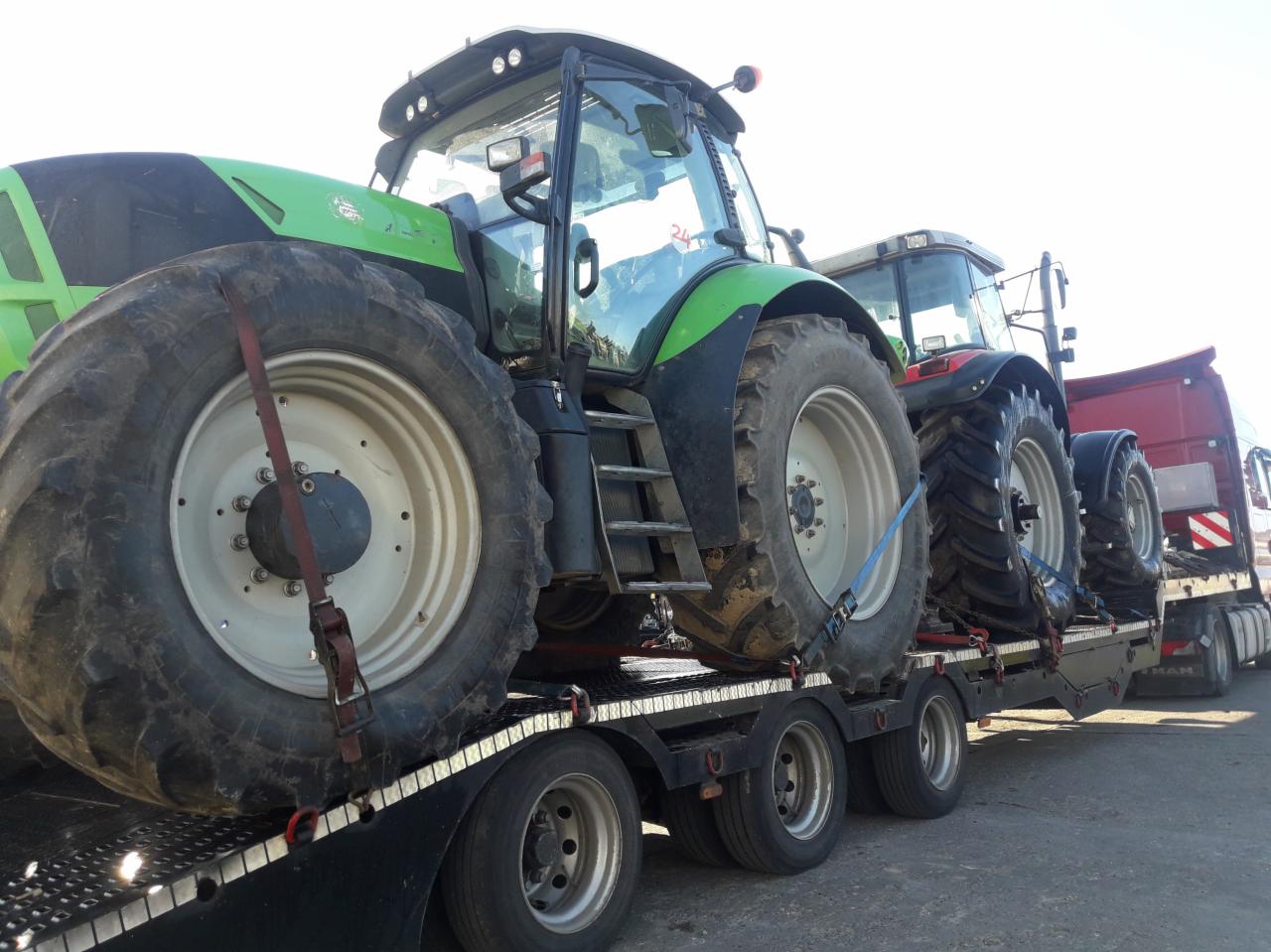 Dwa ciągniki rolnicze, jeden zielony, drugi czerwony, zabezpieczone pasami transportowymi na niskopodwoziowej naczepie ciężarówki, widok z boku na koła i kabiny.