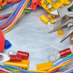 Narzędzia i akcesoria elektryczne: kombinerki z czerwonymi uchwytami, kolorowe przewody, złączki, izolacja i kostki elektryczne na metalowej powierzchni z widocznymi śladami użytkowania.