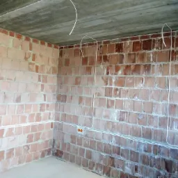 Surowy mur z cegły z widocznymi przewodami elektrycznymi poprowadzonymi w peszlach, w tle betonowy strop.