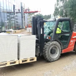 Czerwony wózek widłowy Manitou na budowie, przewożący paletę z płytami gipsowo-kartonowymi; w tle budynek z przeszkloną fasadą i pracownicy na podnośniku.