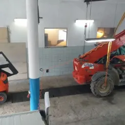 Pomarańczowy wózek widłowy Linde z otwartą kabiną i czerwony podnośnik teleskopowy Maniscopic w hali serwisowej z widocznymi elementami instalacji elektrycznej.