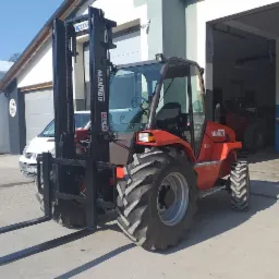 Pomarańczowy wózek widłowy Manitou M26-4 z czarnym masztem na betonowym placu przed budynkiem z otwartą bramą garażową, w tle biały samochód.