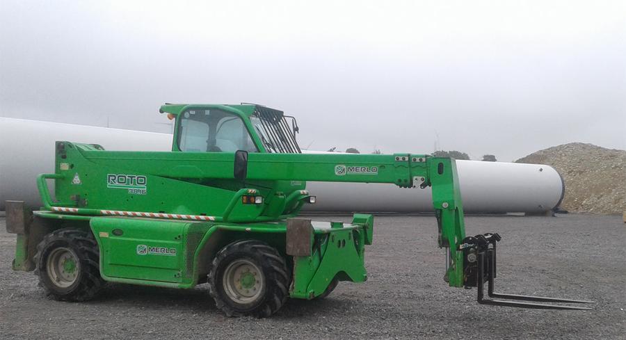 Zielony wózek widłowy teleskopowy Merlo Roto 30.16 na placu budowy, z turbiną wiatrową w tle, pochmurny dzień.