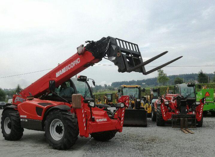 Czerwony teleskopowy wózek widłowy Manitou z podniesionym ramieniem i widłami, na placu z innymi maszynami budowlanymi w tle, pochmurne niebo.