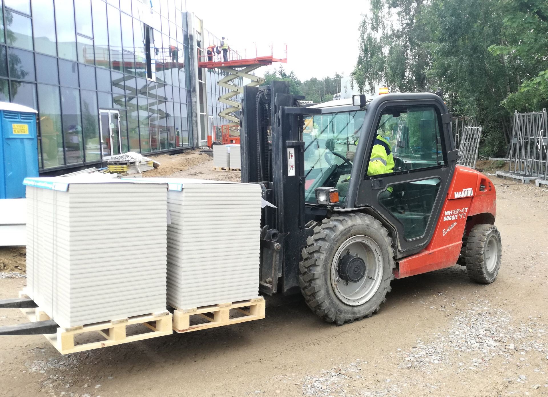 Czerwony wózek widłowy Manitou na budowie, przewożący paletę z płytami gipsowo-kartonowymi; w tle budynek z przeszkloną fasadą i pracownicy na podnośniku.
