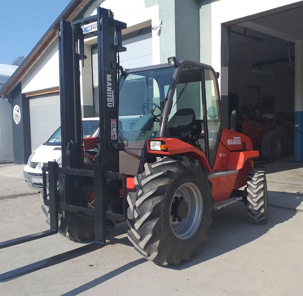 Pomarańczowy wózek widłowy Manitou M26-4 z czarnym masztem na betonowym placu przed budynkiem z otwartą bramą garażową, w tle biały samochód.