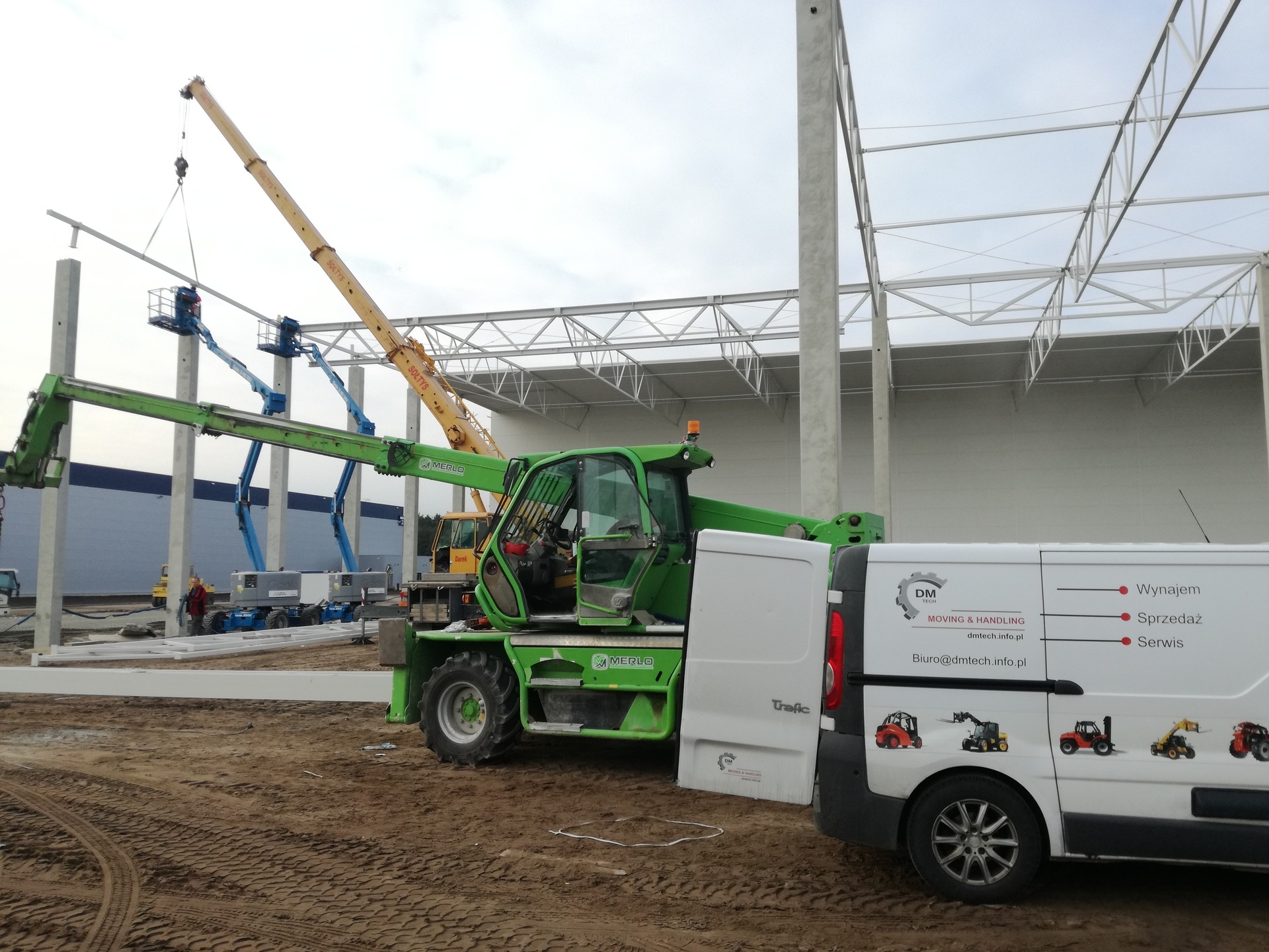 Montaż konstrukcji stalowej hali: zielony teleskopowy wózek widłowy Merlo, żuraw Soltys i podnośniki koszowe w akcji, na tle niedokończonej hali i zaparkowanego busa z reklamą firmy.