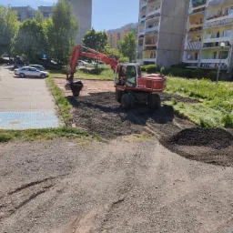 Czerwona koparka Atlas 1404 z operatorem wyrównuje teren podłoża w otoczeniu bloków mieszkalnych i drzew, przygotowując obszar pod przyszłą inwestycję.