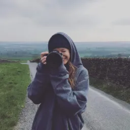 Uśmiechnięta kobieta w szarej bluzie z kapturem i czapce, trzyma aparat Sony na tle krajobrazu z kamiennym murem i drogą.