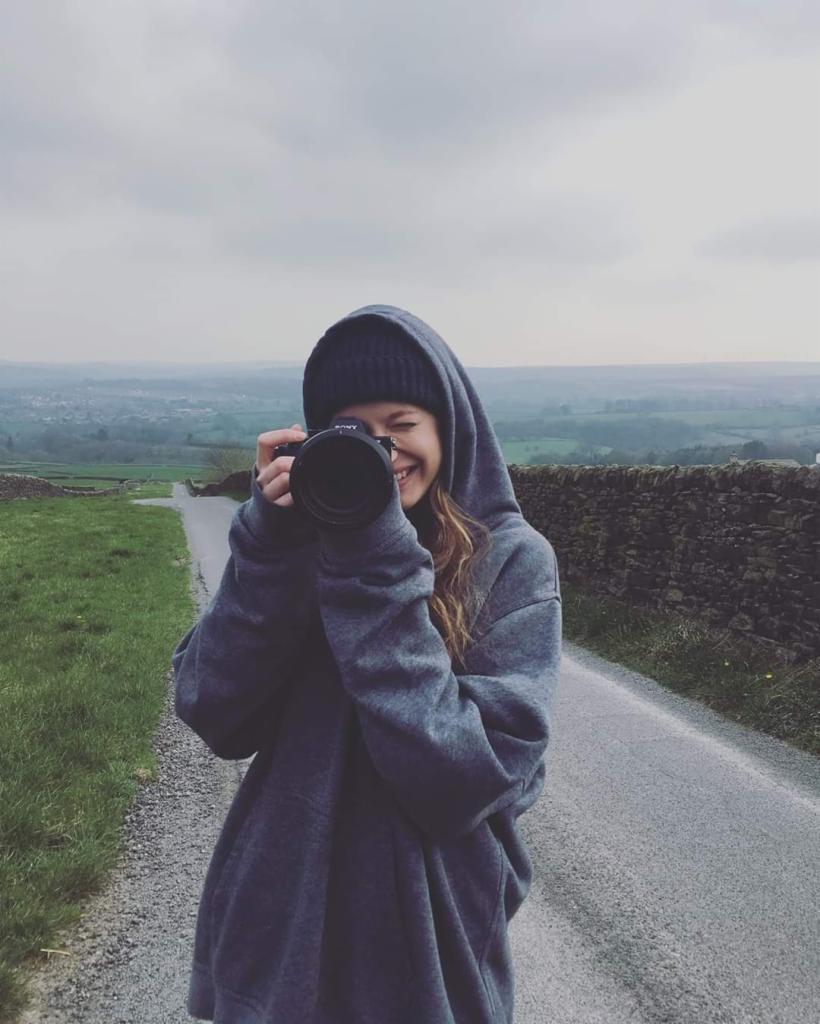 Uśmiechnięta kobieta w szarej bluzie z kapturem i czapce, trzyma aparat Sony na tle krajobrazu z kamiennym murem i drogą.
