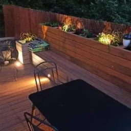 Drewniany taras z podwyższonymi rabatami, oświetleniem punktowym, dwoma ażurowymi fotelami i czarnym stołem, oświetlony wieczorową porą.